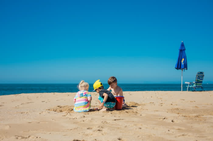 Three children digging in the sand with a bucket, beside a beach umbrella and chair